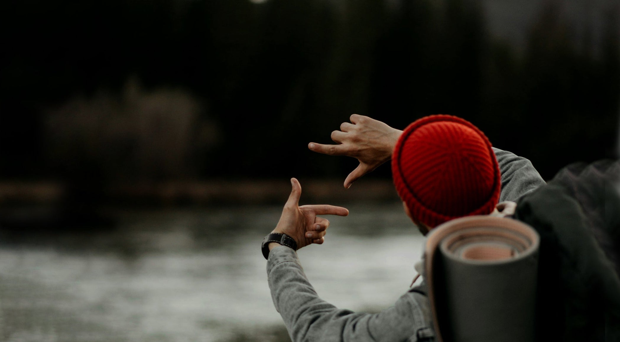 Person in a red beanie and gray jacket pointing towards a body of water with a backpack and thermos.